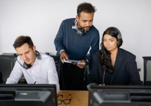 What is a line manager? Friendly, smiling male manager wearing a jumper looking over shoulder of two employees sat at computers