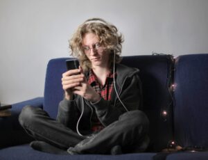 How to overcome communication barriers. Young man with long hair sat on sofa looking at his phone, wearing headphones.