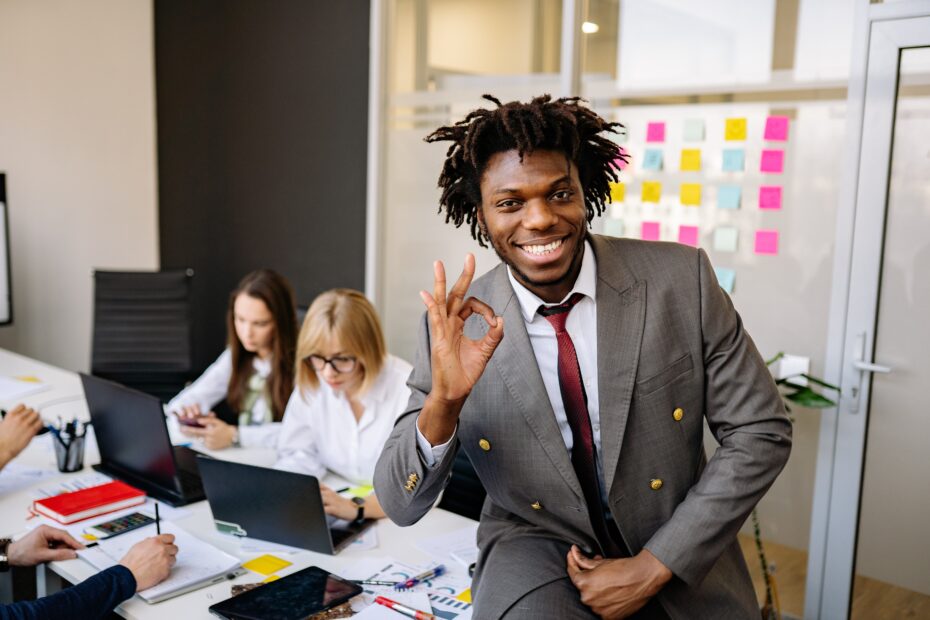 How to be a good team leader. Young man in suit smiling and waving at the camera with two young women sat looking at laptops in the background.