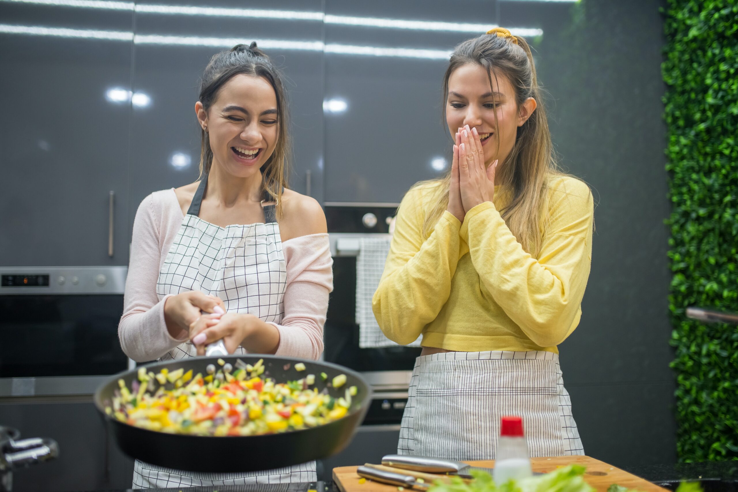 Fire safety quiz - kitchen safety. Two young women smiling whilst one woman tosses vegetables in a frying pan.