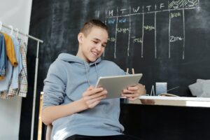 Money worksheets - budgeting. Male teenager sat in school holding an ipad, wearing a blue hoodie.